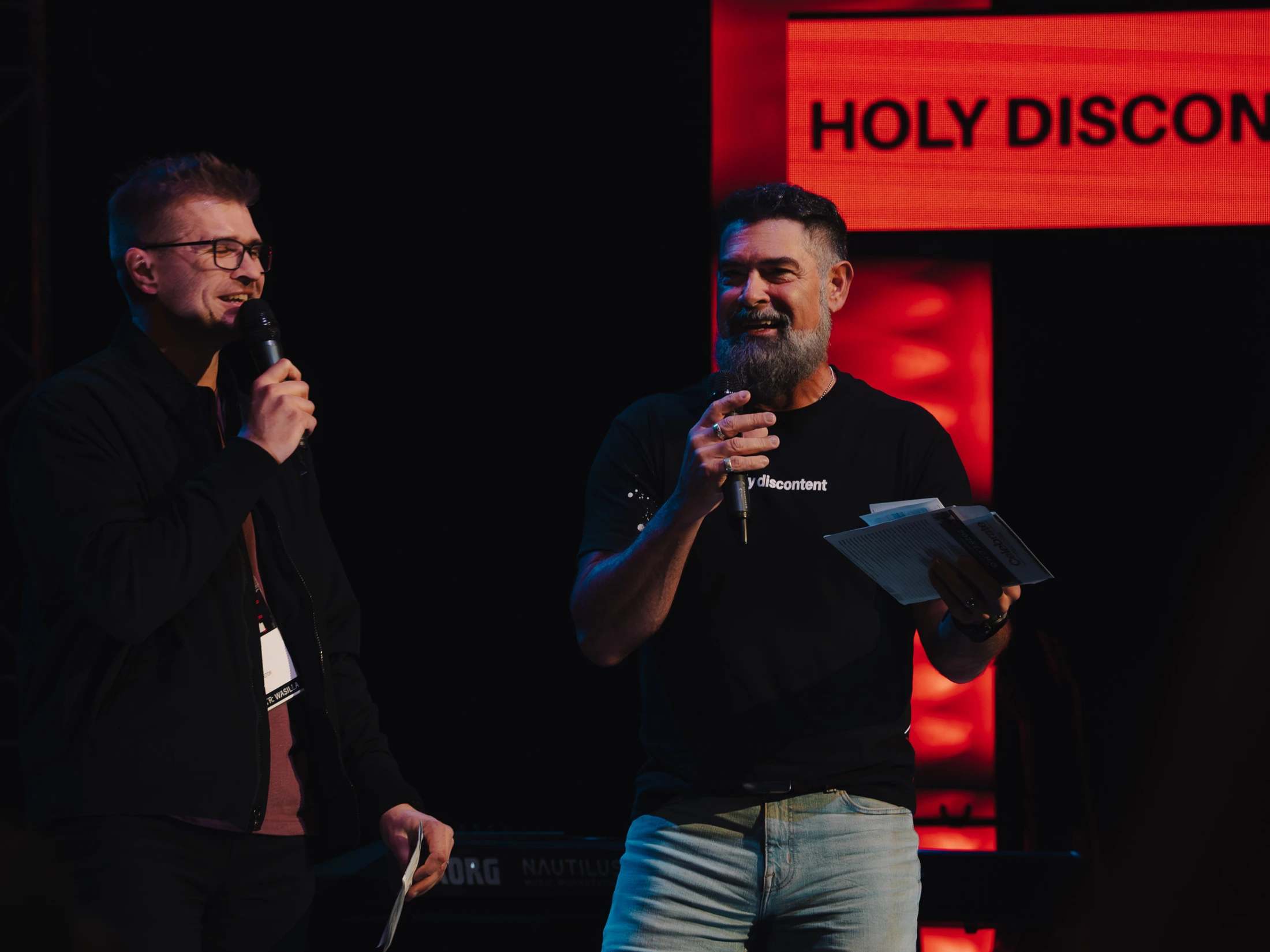 Two men stand on a dimly lit stage, smiling and speaking into microphones. Behind them, a large red screen displays the words 'HOLY DISCONTENT'.